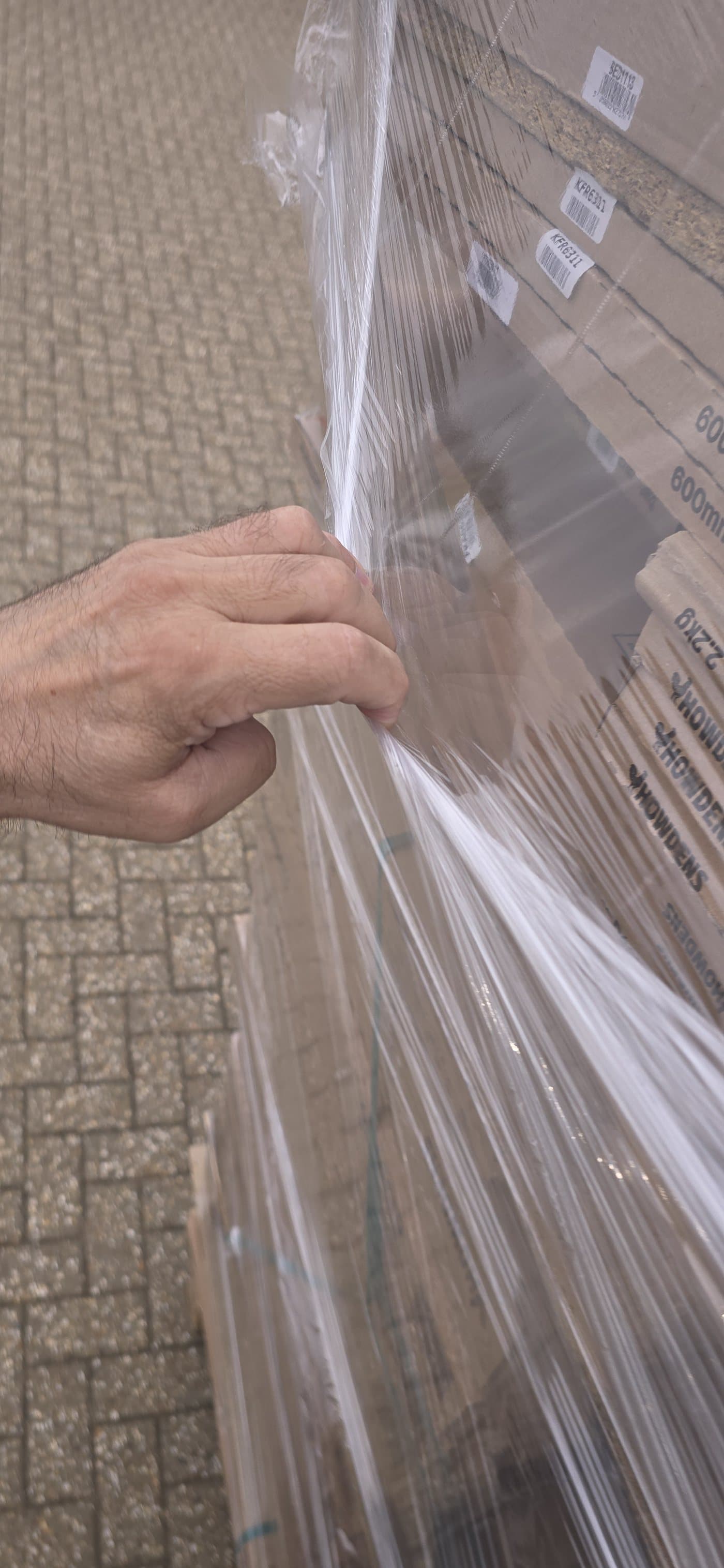 A hand gripping the applied Hyper Wrap film at the side of the load and pulling it away — the film stretches instead of tearing
